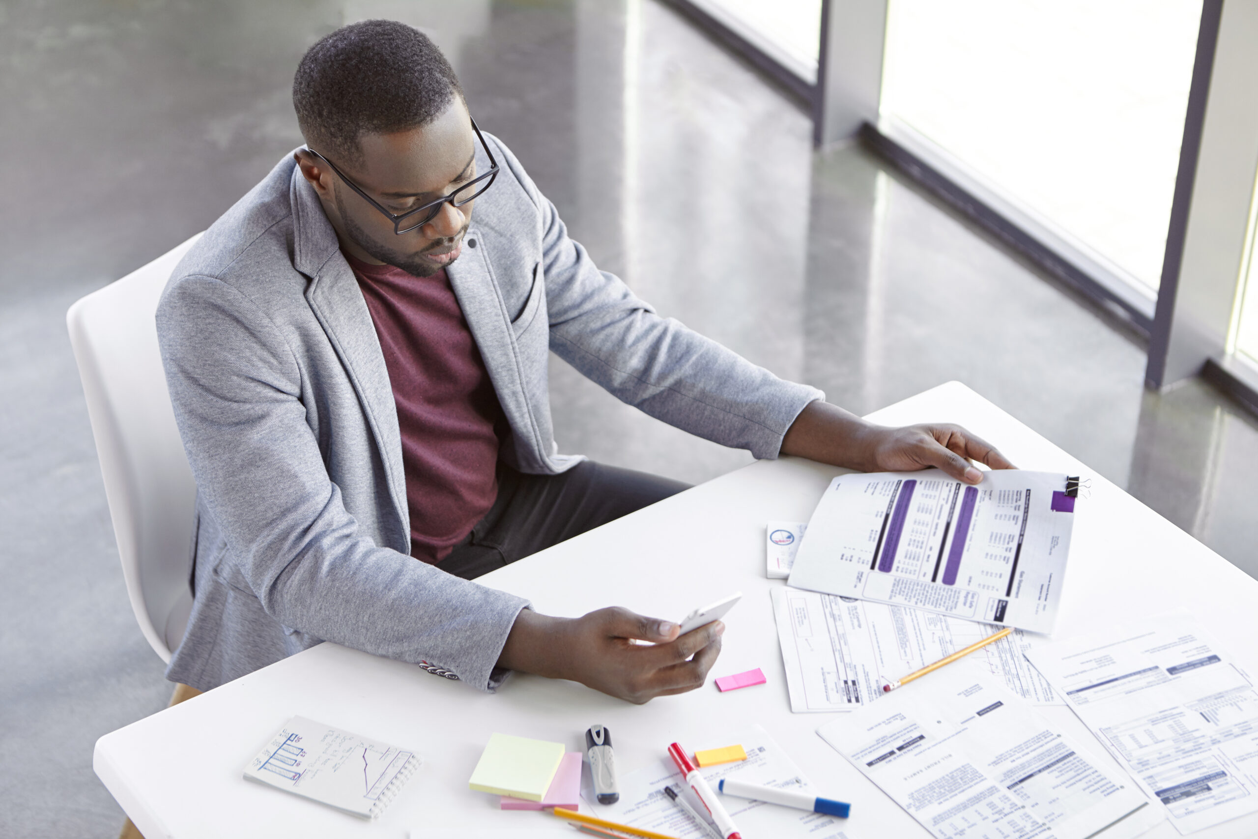 Portrait of successful prosperus dark skinned male works with documents, searches important information on websites, uses modern mobile phone and free internet connection in office, types text